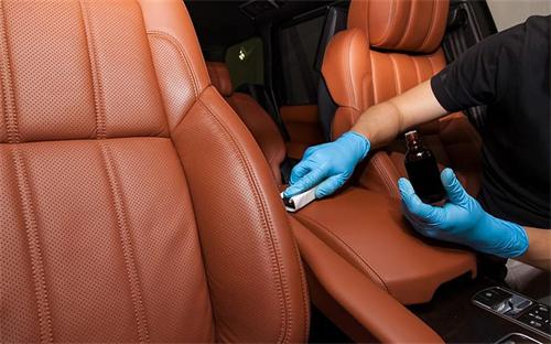 A worker wearing blue protective gloves cleaning brown perforated leather car seats with maintenance supplies