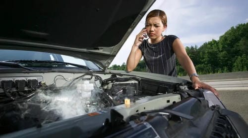 "A woman making a roadside assistance phone call beside her overheating car with steam rising from the engine bay"