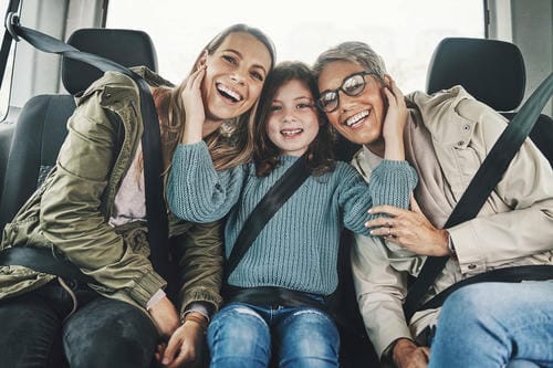 "Three smiling female family members wearing seat belts sitting together on a car rear seat"
