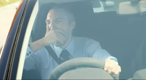 "Tired male driver wearing a seatbelt yawning while sitting behind the steering wheel inside a car"