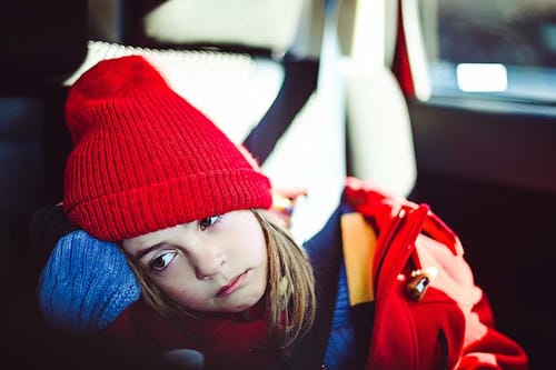 "Fatigued little girl wearing a red knit hat resting her head against her arm in the car rear seat, feeling carsick and listless"