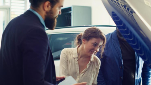 The auto sales consultant introduces the spare tire configuration and vehicle condition to a couple of customers beside the open car trunk.