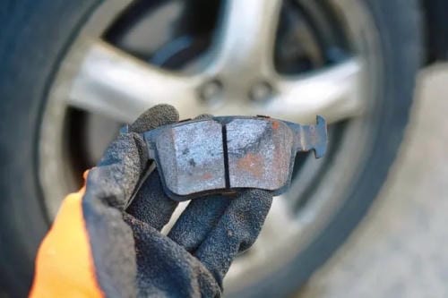 "Mechanic's gloved hand holding a worn old car brake pad in front of a vehicle alloy wheel"