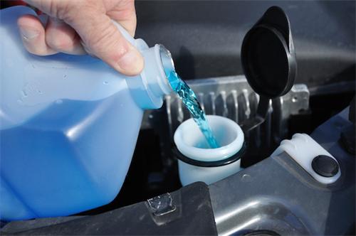 "Driver pouring blue antifreeze windshield washer fluid into the car's engine bay reservoir"